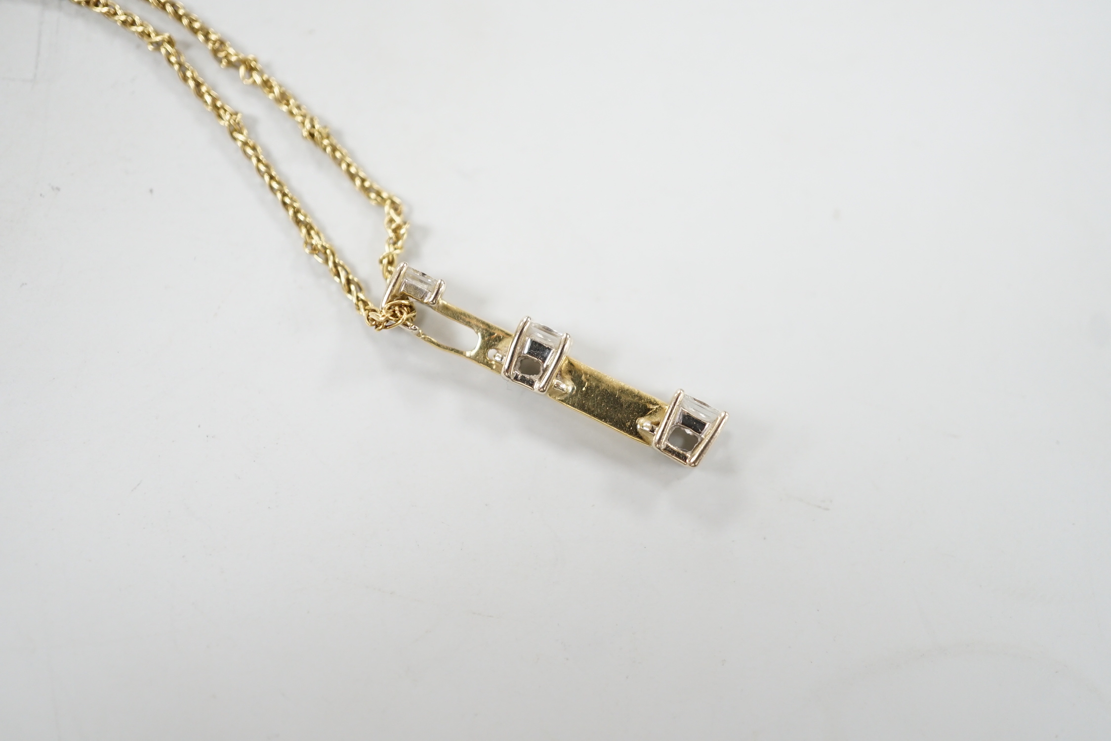 A modern yellow metal and graduated three stone princess cut diamond set line pendant, 25mm, on a 750 chain, 38cm, gross weight 4.5 grams.